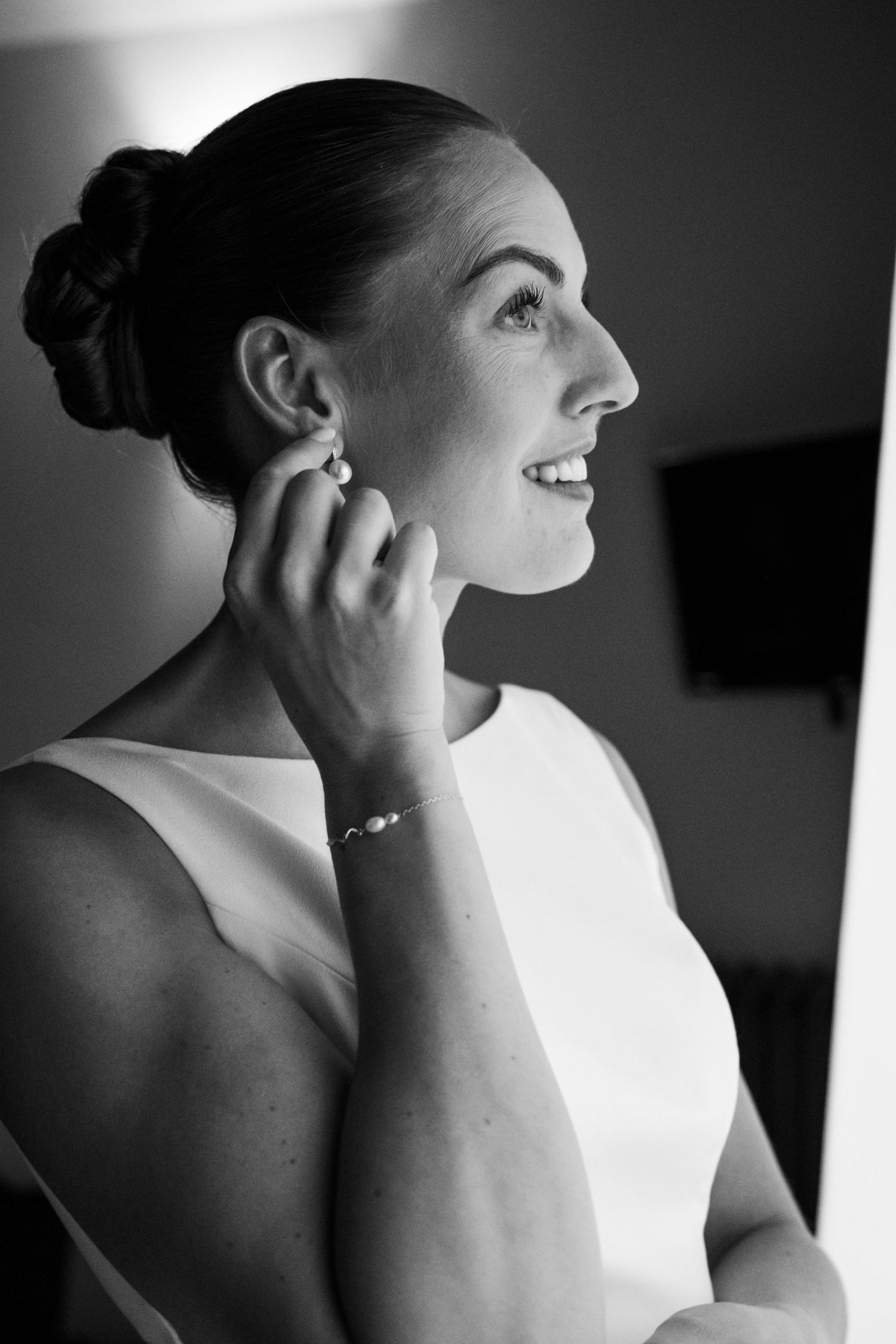 Black and white photo of a smiling bride putting on pearl earrings, wearing a sleeveless white dress and bracelet.