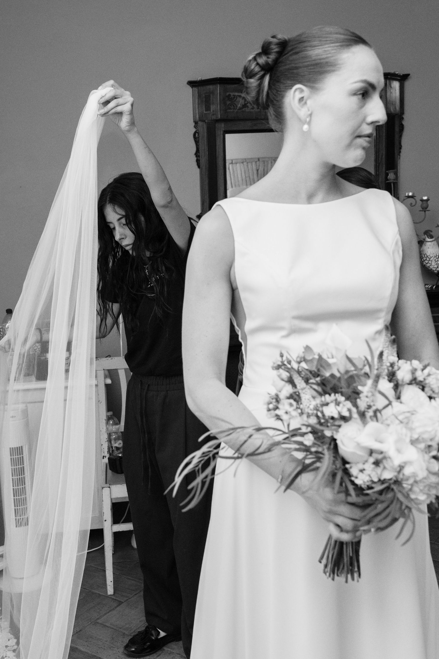 Black and white photo of a bride holding a bouquet while a bridesmaid adjusts her veil indoors.