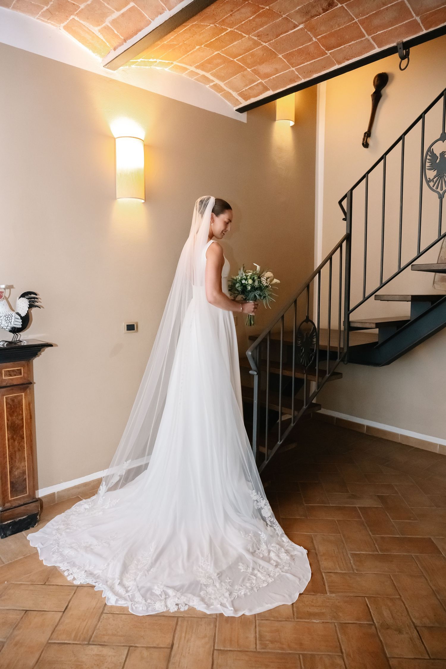 Bride in a white gown with long veil holds bouquet near spiral staircase in elegant villa interior.