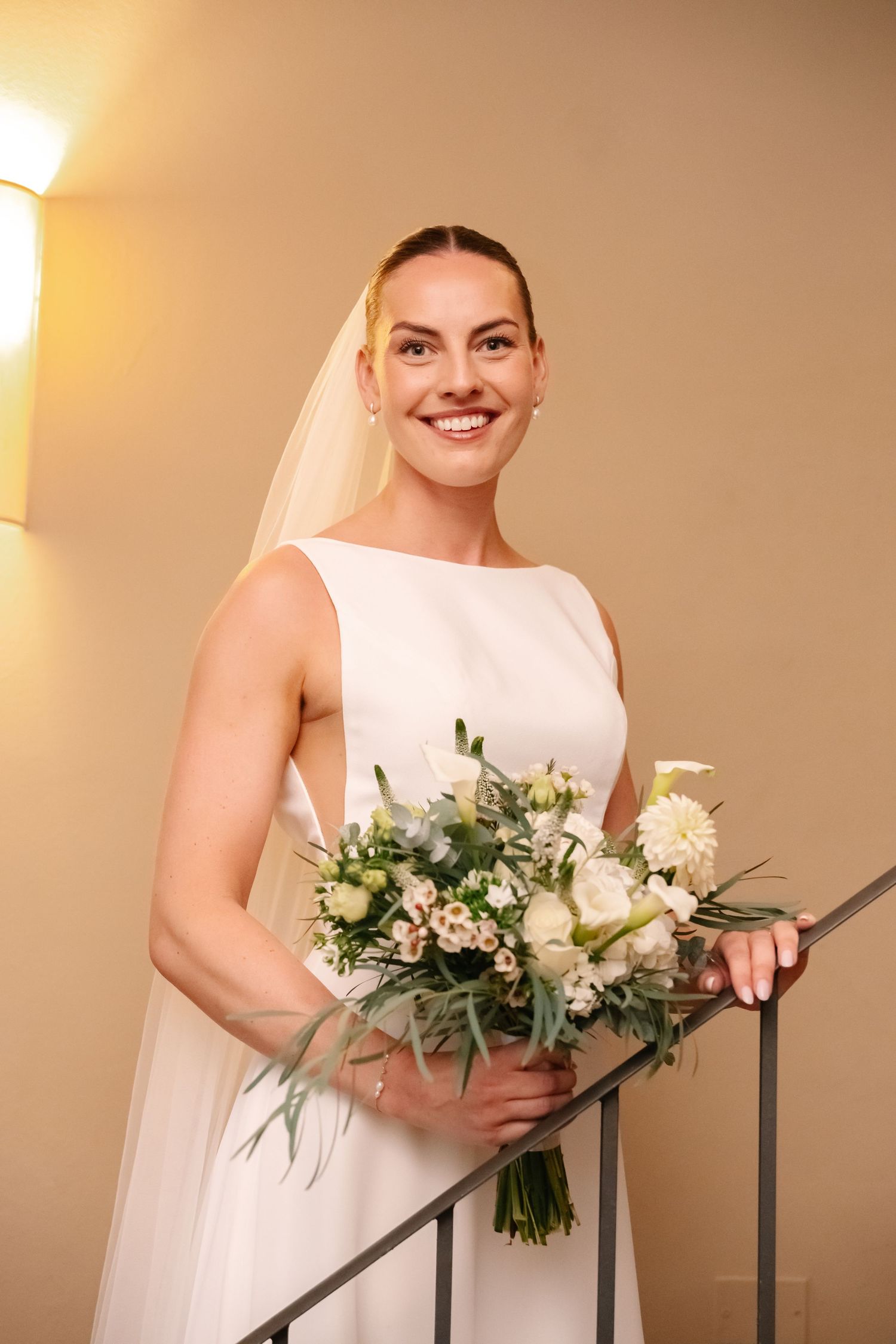 Smiling bride in white sleeveless gown and veil holding a bouquet of white flowers on a staircase.