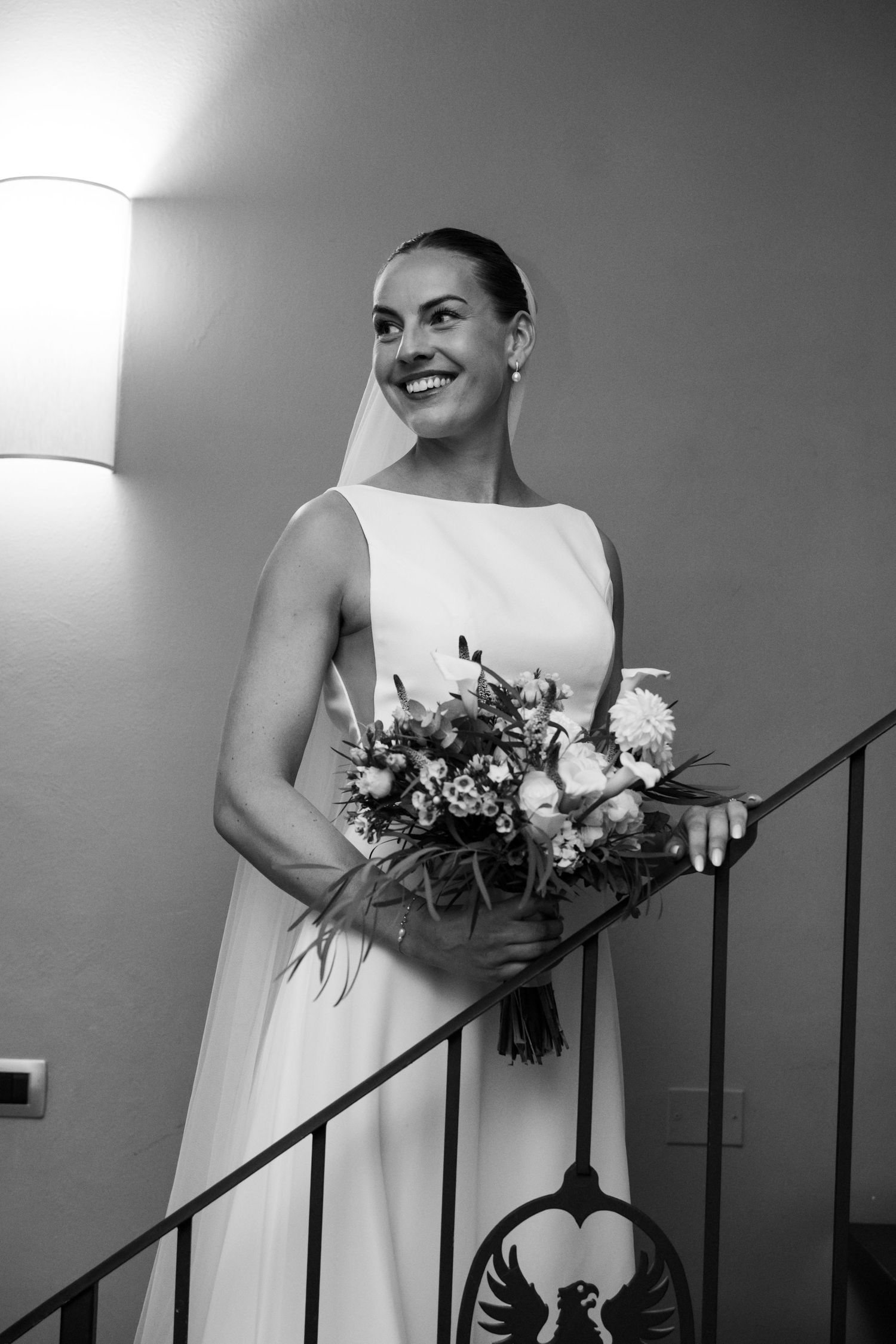 Smiling bride in white gown holding bouquet on staircase, black and white wedding photo.