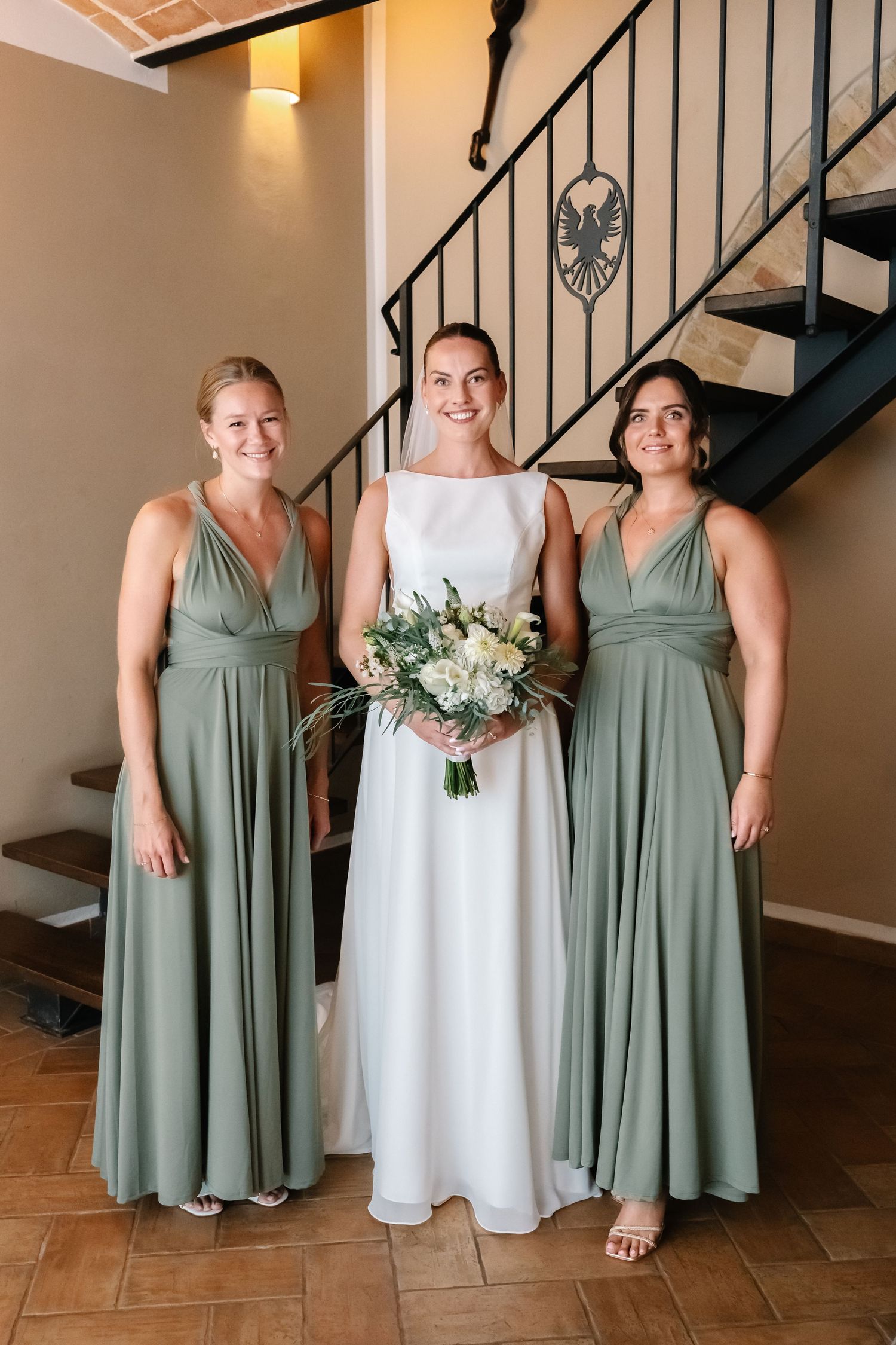 Bride in white gown holds bouquet flanked by two bridesmaids in sage green dresses near staircase.