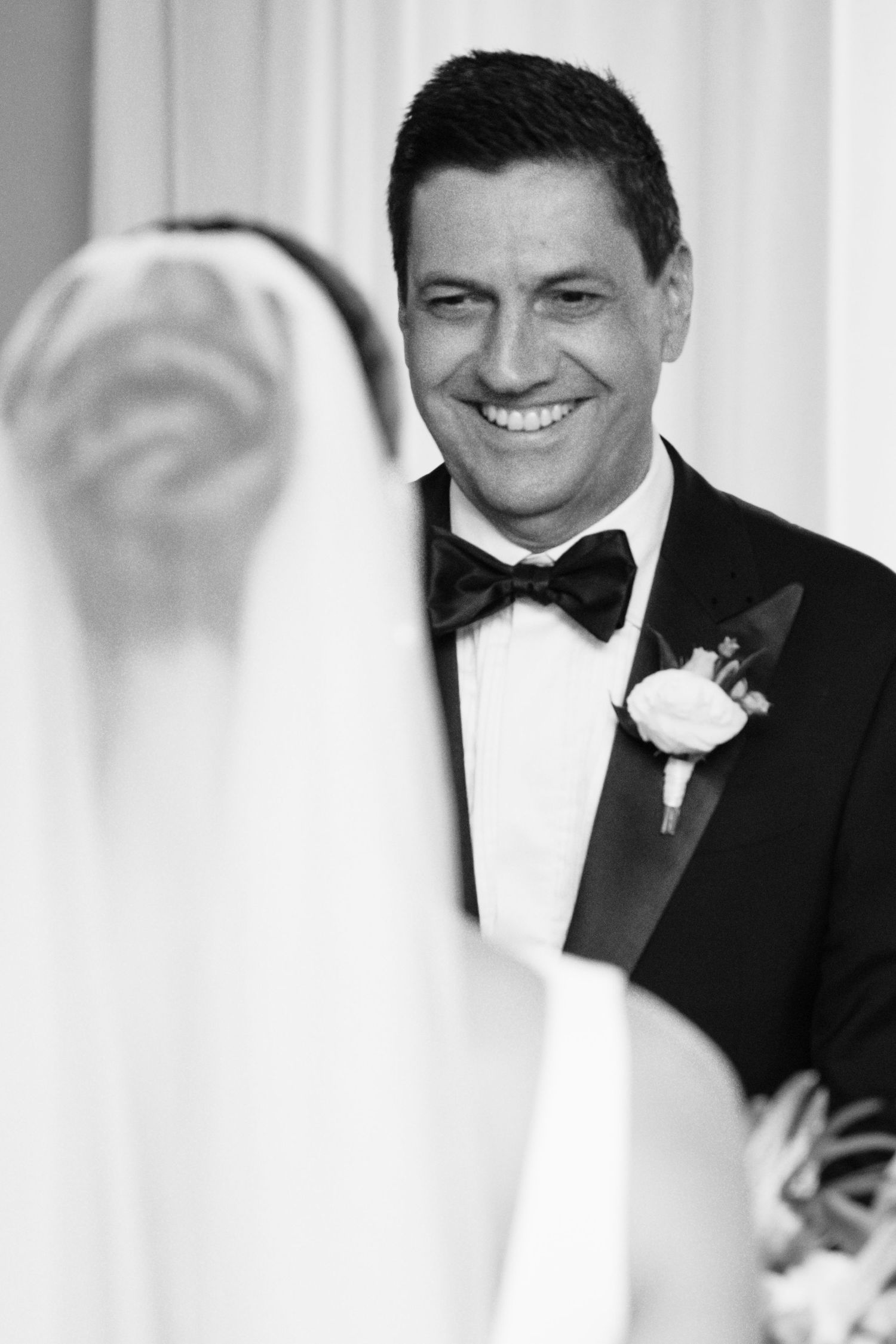 Groom in black tuxedo smiling at bride in veil during wedding ceremony, black and white photo.