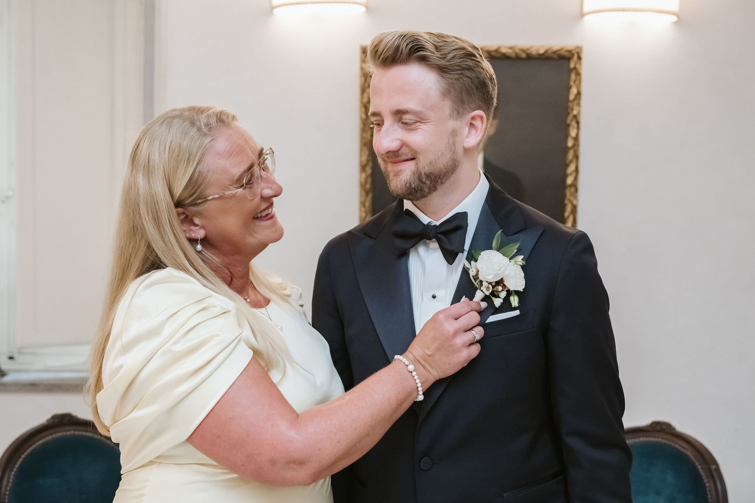 A groom in a tuxedo smiles as an older woman pins a boutonniere on his lapel at a wedding ceremony.