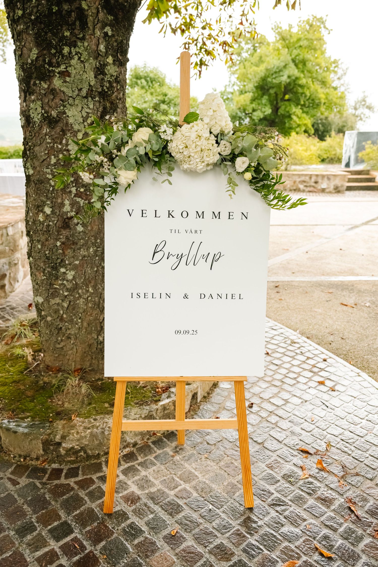 Elegant wedding welcome sign on easel reading Velkommen Til Vart Bryllup, decorated with white hydrangeas and greenery.