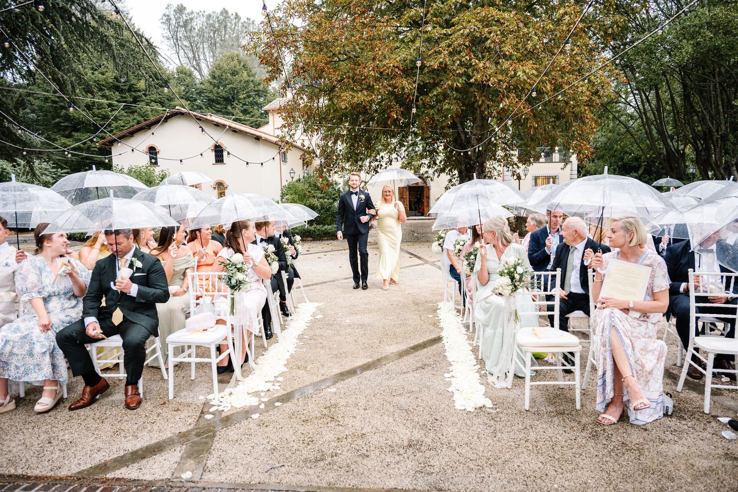 Outdoor wedding ceremony with guests holding clear umbrellas seated on white chairs along a petal-lined aisle.