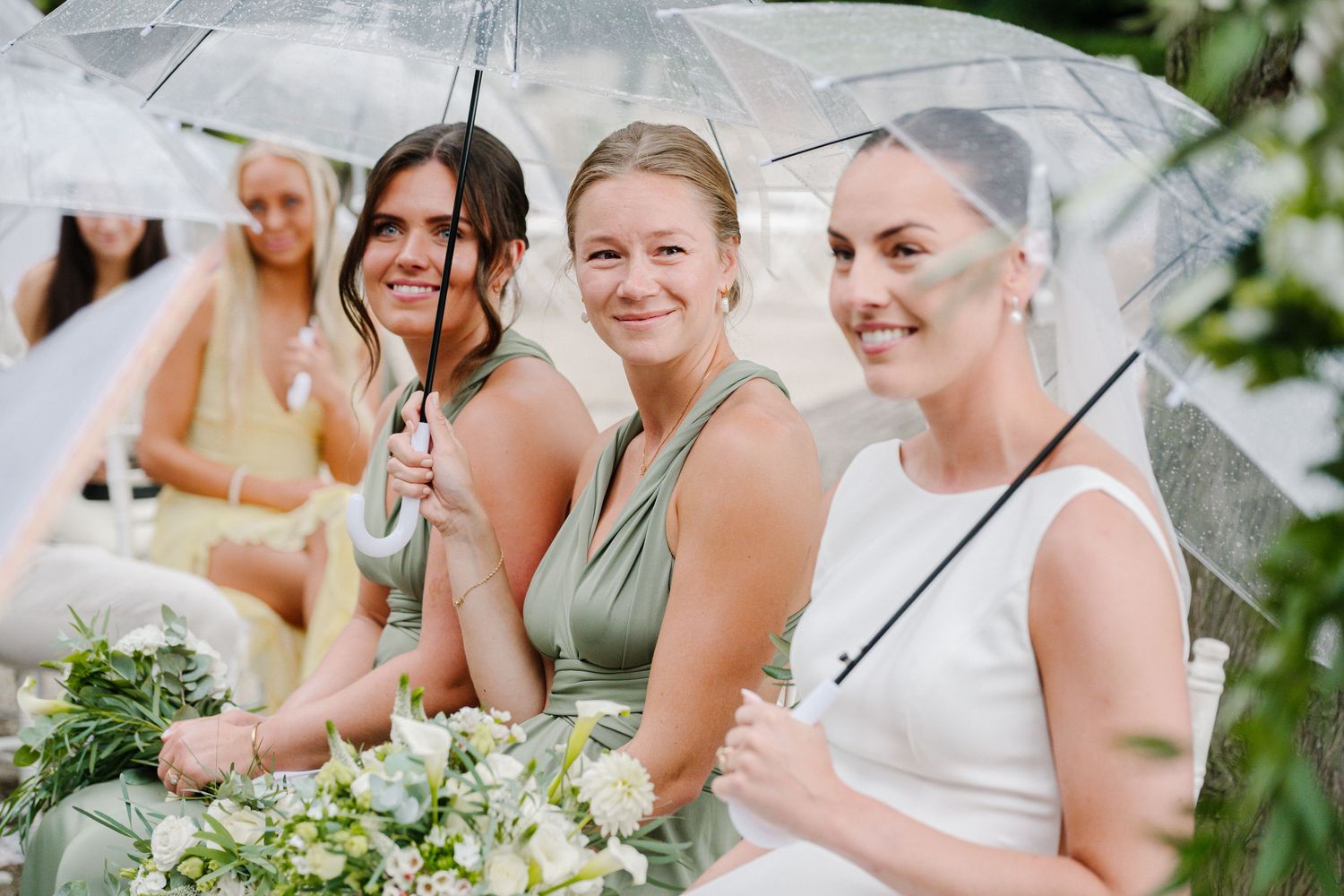Three bridesmaids smiling under clear umbrellas at an outdoor wedding ceremony, holding white floral bouquets.