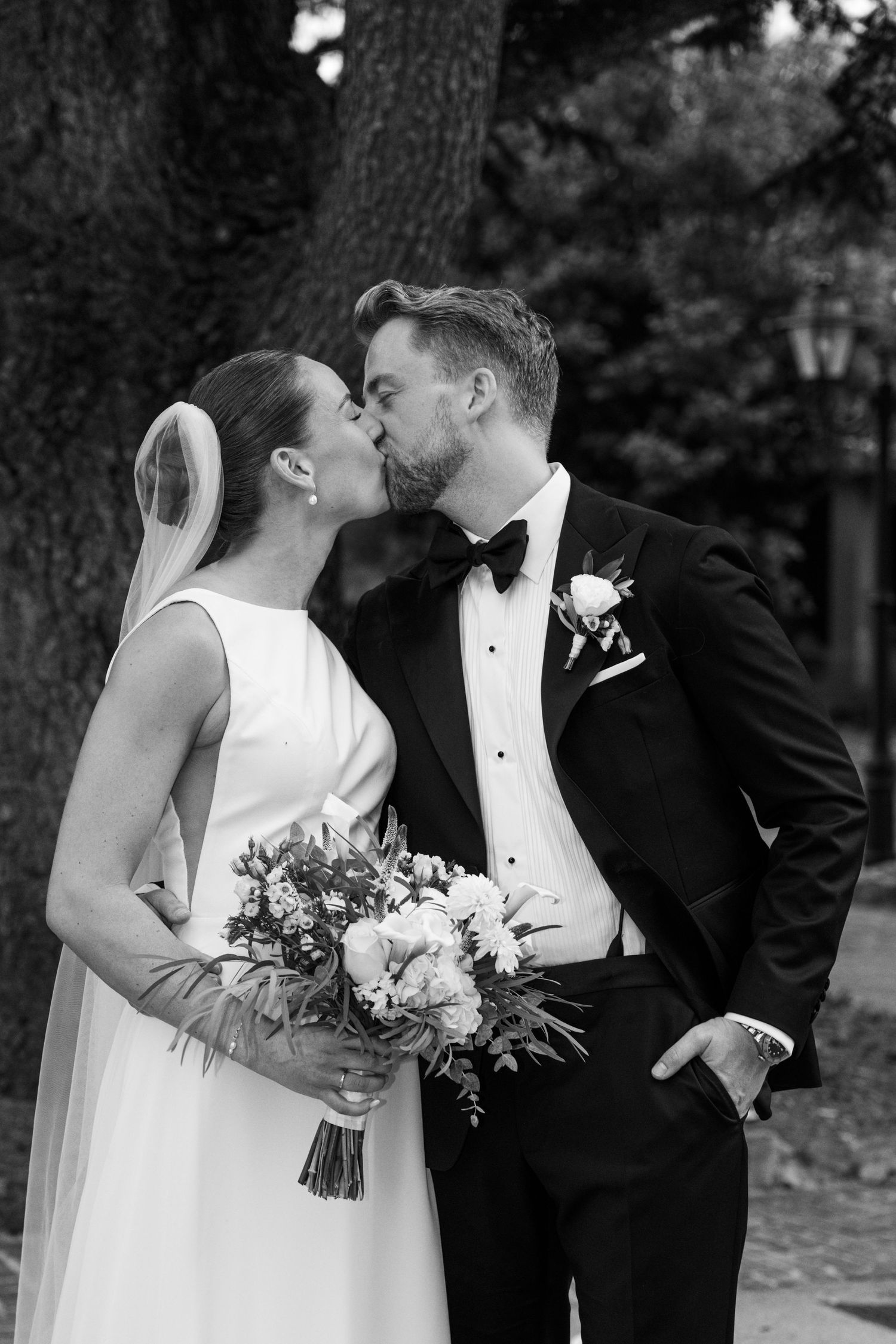 Black and white photo of a bride and groom sharing a kiss outdoors, bride holding a bouquet in a white gown.