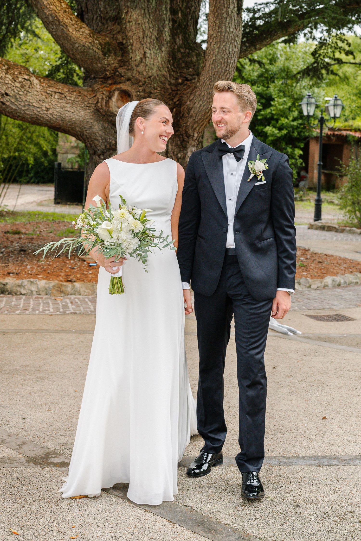 Bride and groom smiling at each other outdoors, she holds a white bouquet, he wears a black tuxedo with boutonniere.