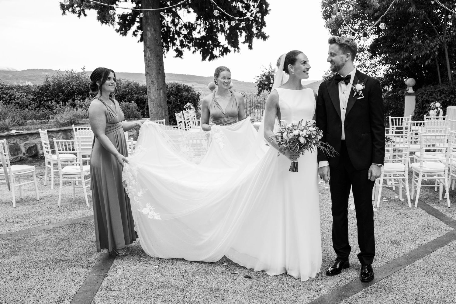 Black and white wedding photo of bride and groom with two bridesmaids adjusting bridal gown outdoors.