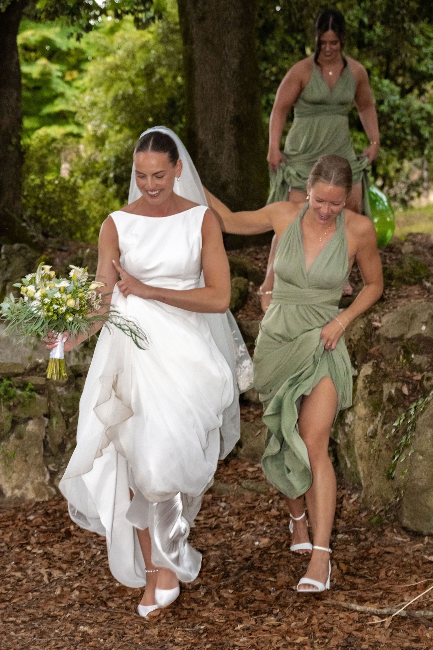 Bride in white gown with bridesmaids in sage green dresses walking through a garden wedding venue.