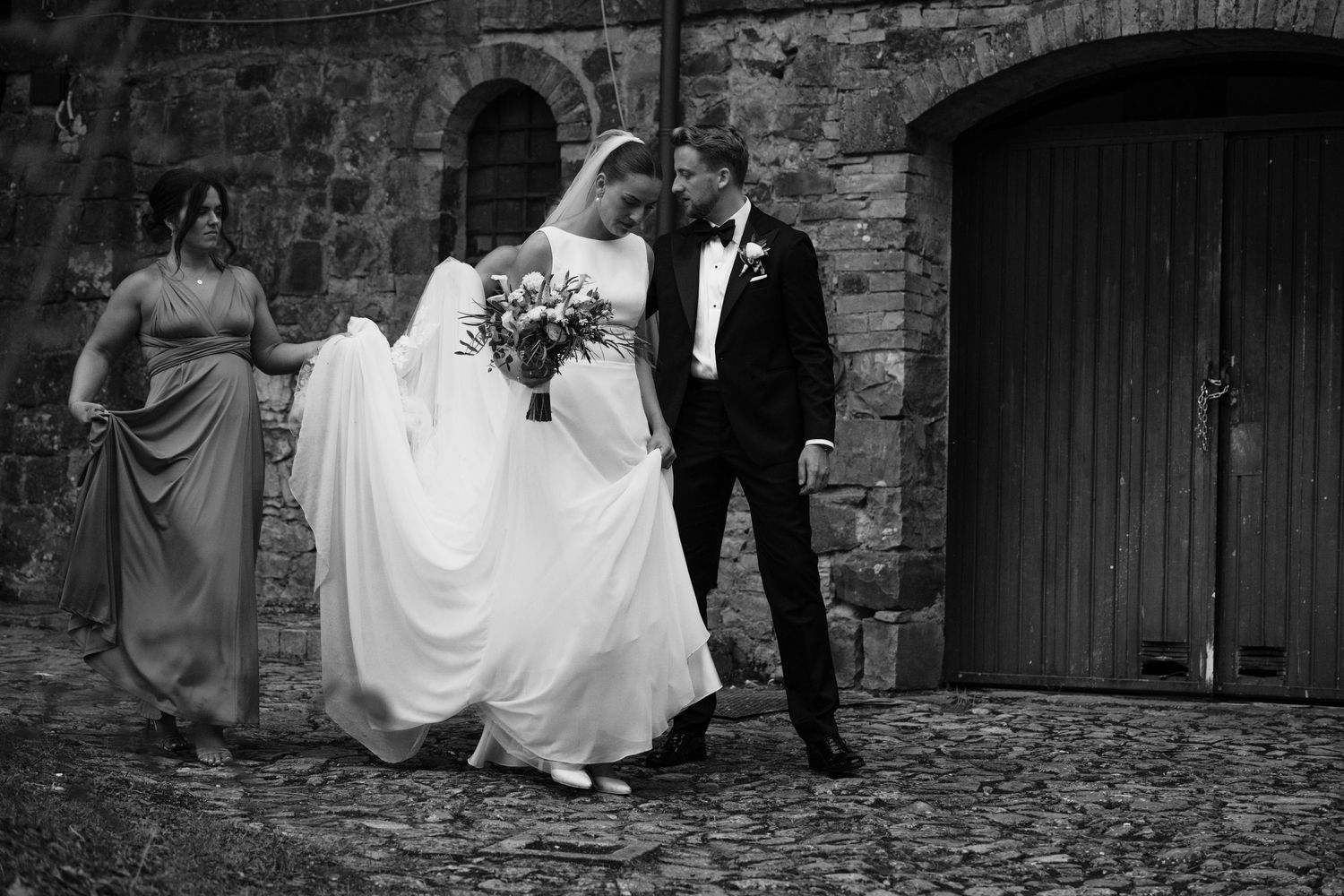 Black and white wedding photo of bride and groom walking with bridesmaid near stone wall.