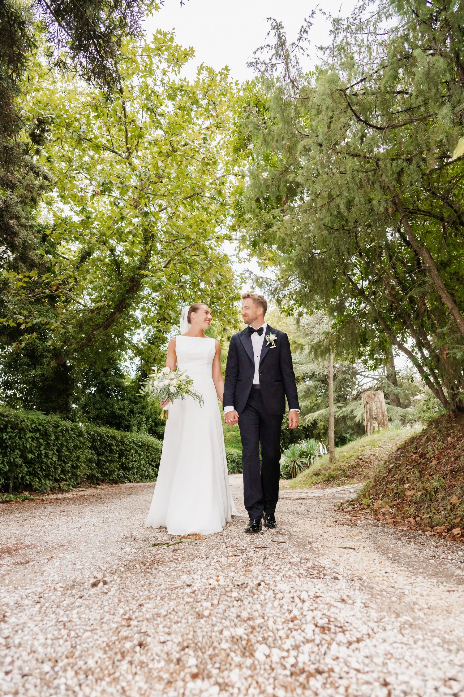 Bride and groom walking hand in hand on a gravel path surrounded by lush green trees on their wedding day.