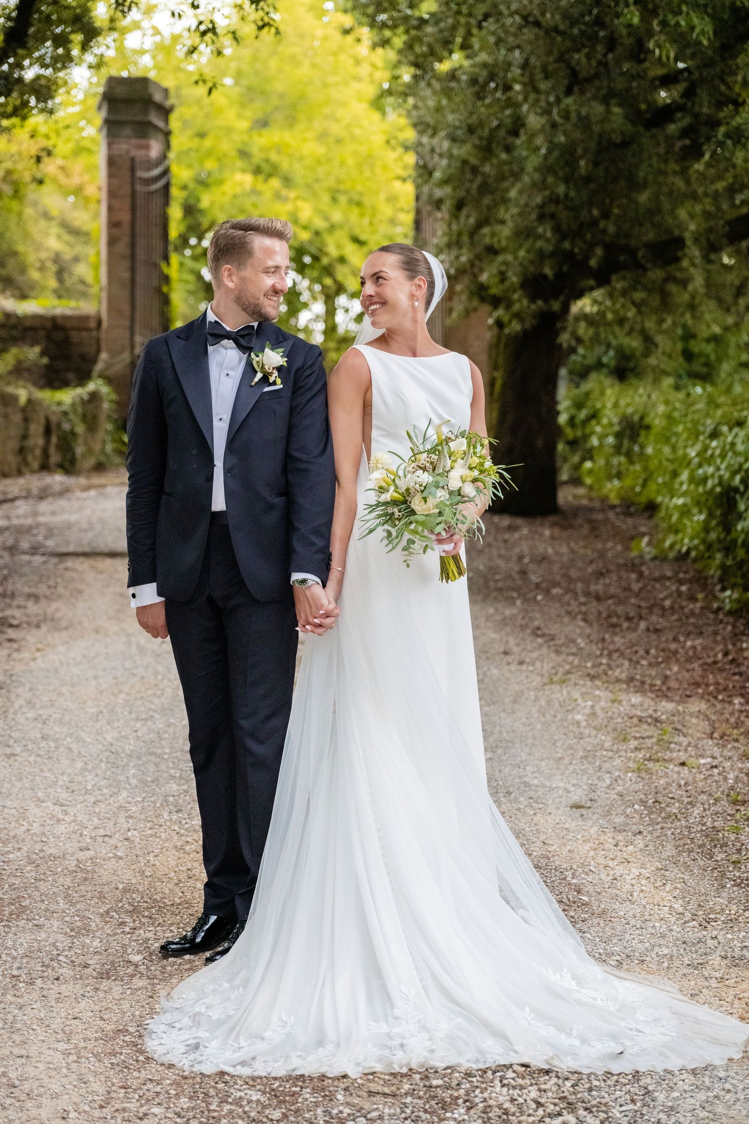 Bride and groom holding hands on a gravel path, smiling at each other, with lush green trees in the background.