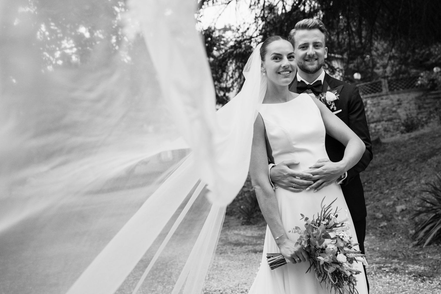 Black and white wedding photo of smiling bride in gown with flowing veil and groom in suit holding bouquet outdoors.