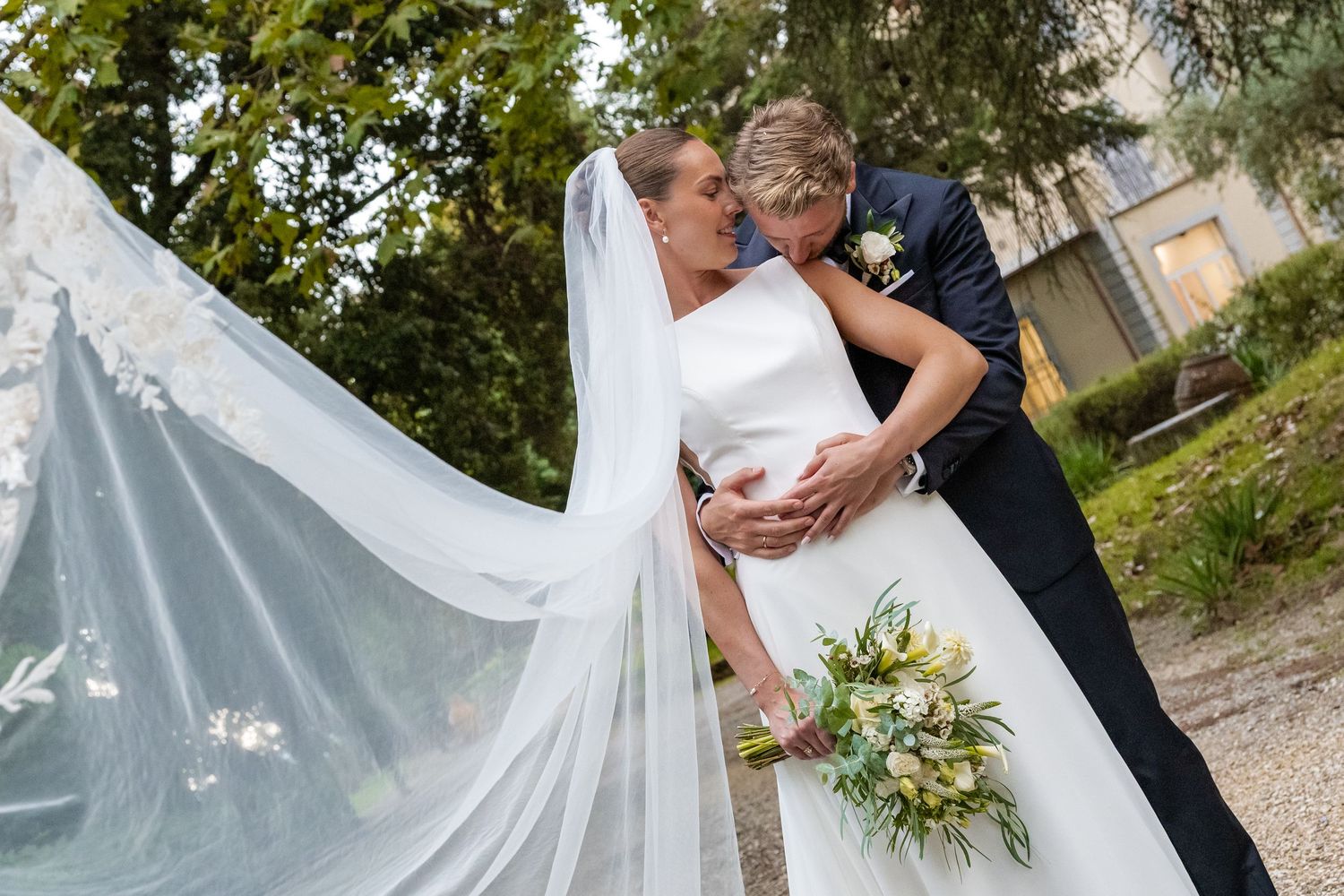 Bride and groom sharing a romantic kiss outdoors, with flowing white veil and bridal bouquet on their wedding day.