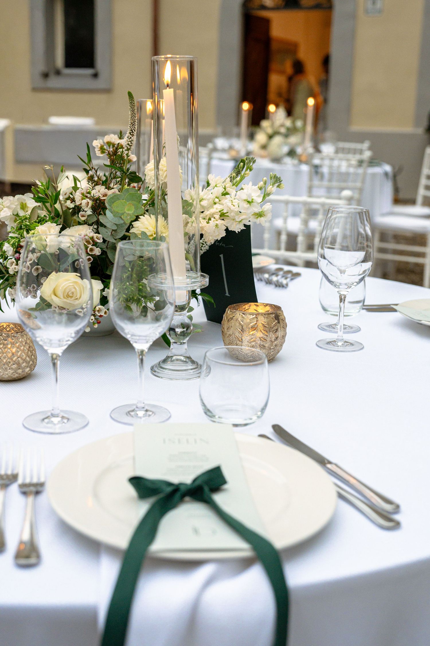 Elegant wedding reception table with white florals, tall candles, crystal glasses, and green ribbon-adorned place settings.