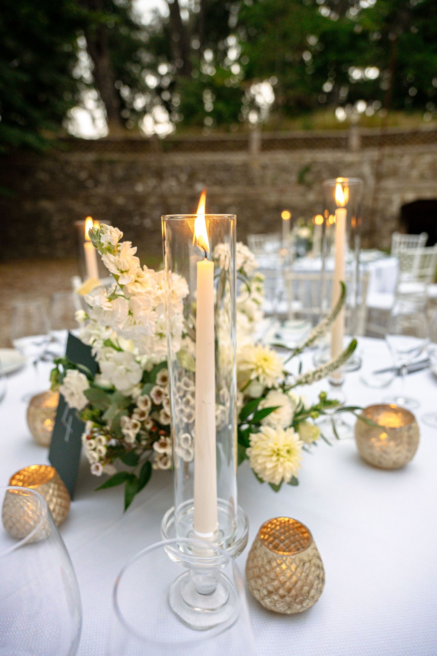 Elegant wedding table centerpiece with tall white candles, white floral arrangement, and gold votive candles on white linen.