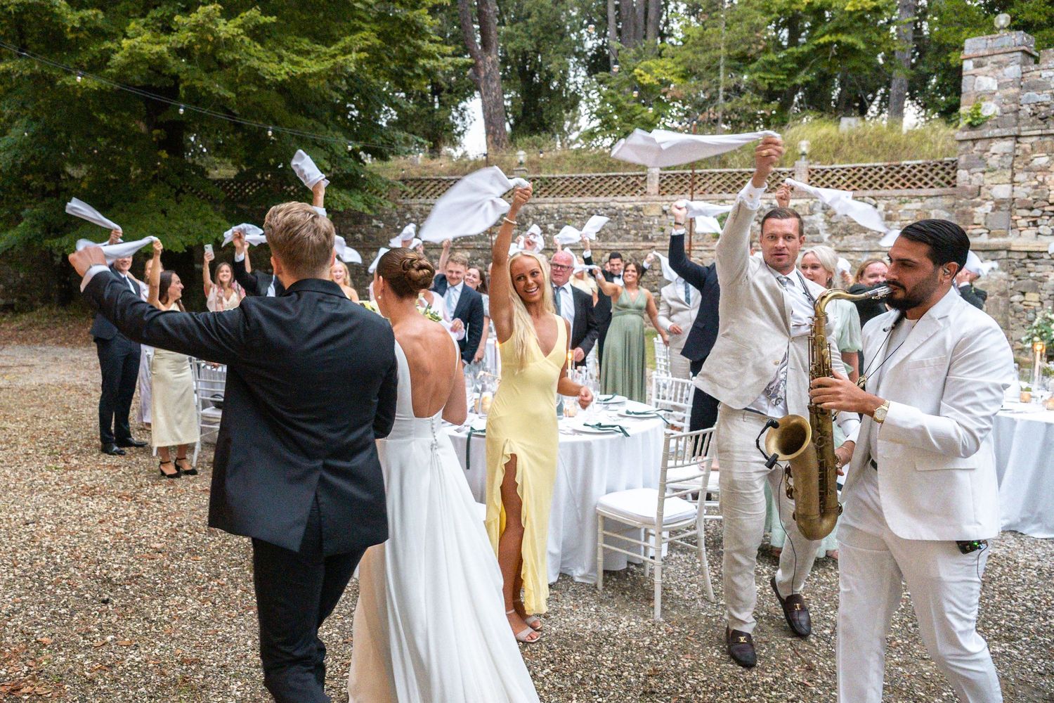 Joyful wedding celebration outdoors with guests cheering, bride in white gown, and saxophonist performing at reception.