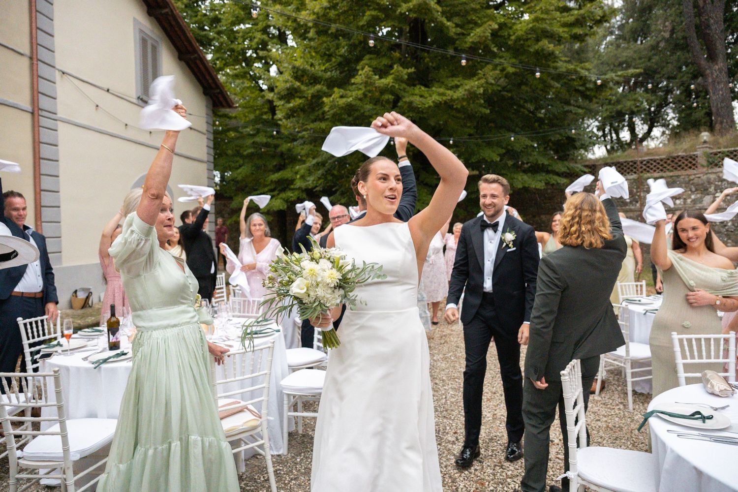 Bride waves white paper at outdoor wedding celebration with guests and floral arrangements at elegant venue.