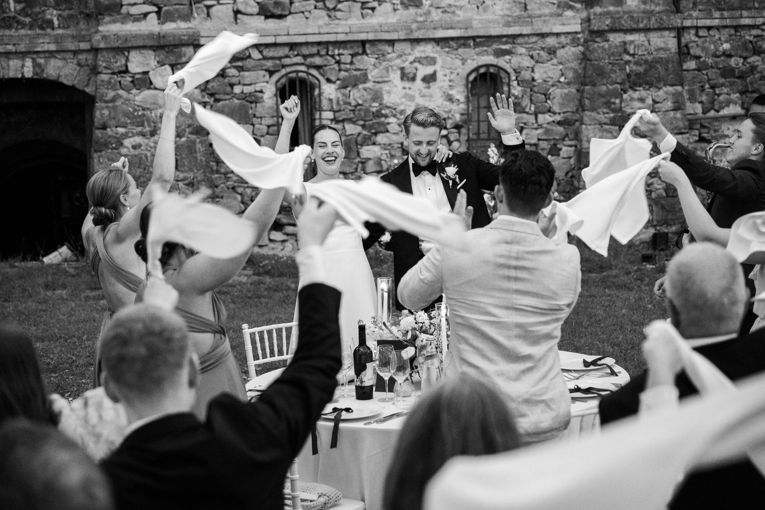 Black and white photo of bride and groom celebrating with guests waving napkins at an outdoor wedding reception.