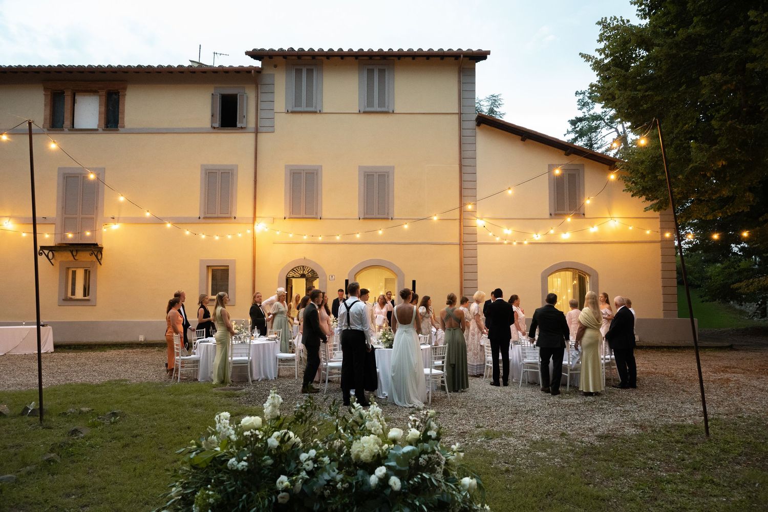 Elegant outdoor wedding reception at a Tuscan villa with string lights, guests mingling, and floral arrangements at dusk.