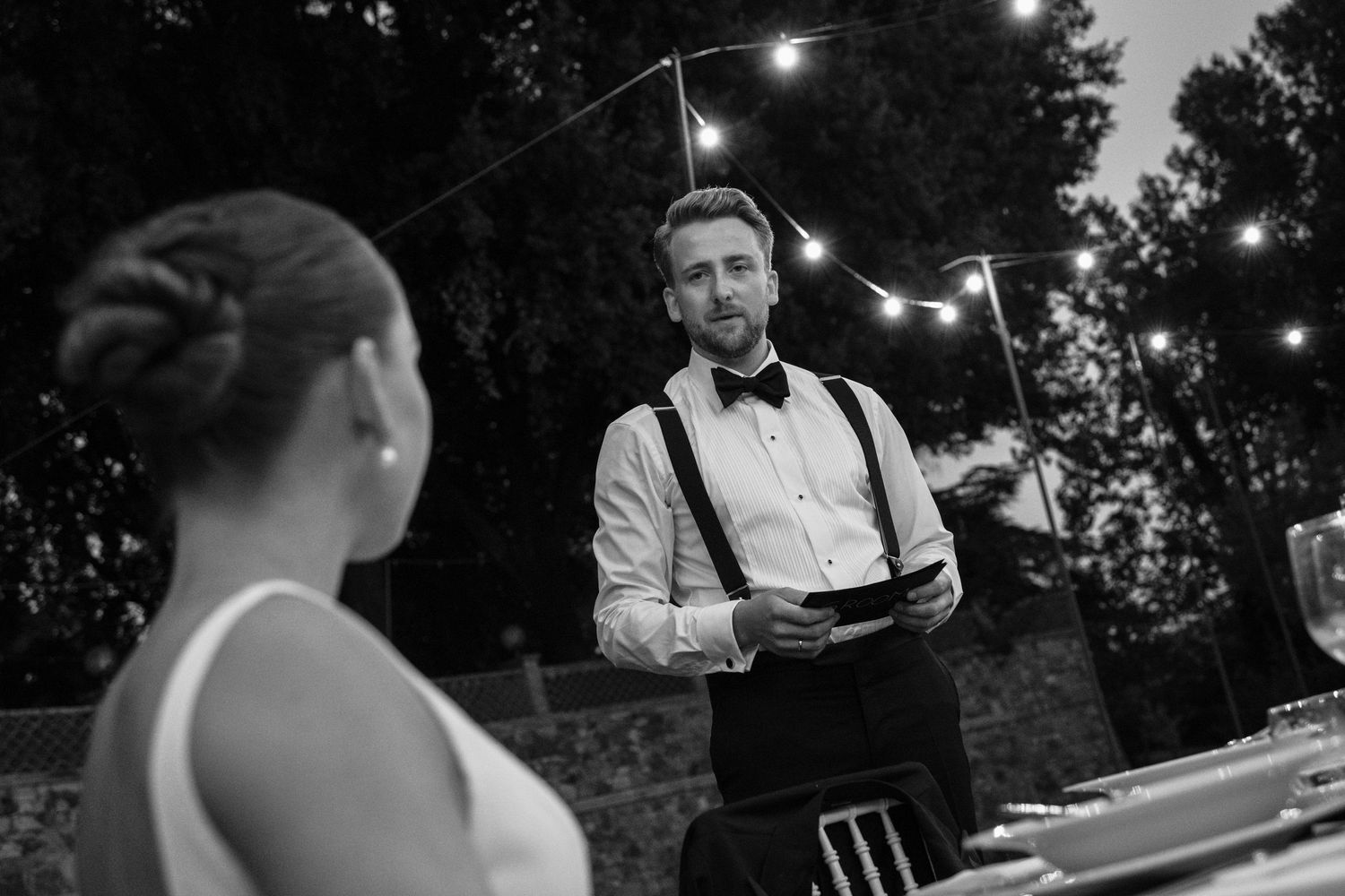 Black and white wedding photo of groom in bow tie and suspenders speaking to bride outdoors with string lights.