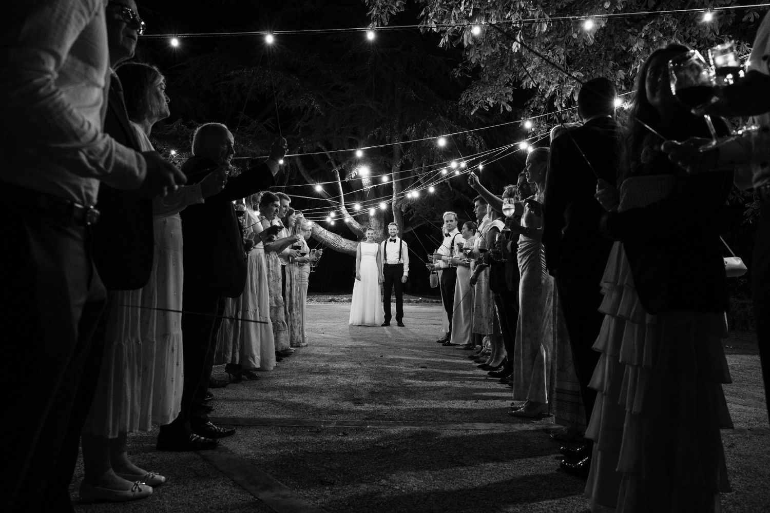 Black and white wedding send-off with guests lining a path under string lights as couple walks through.
