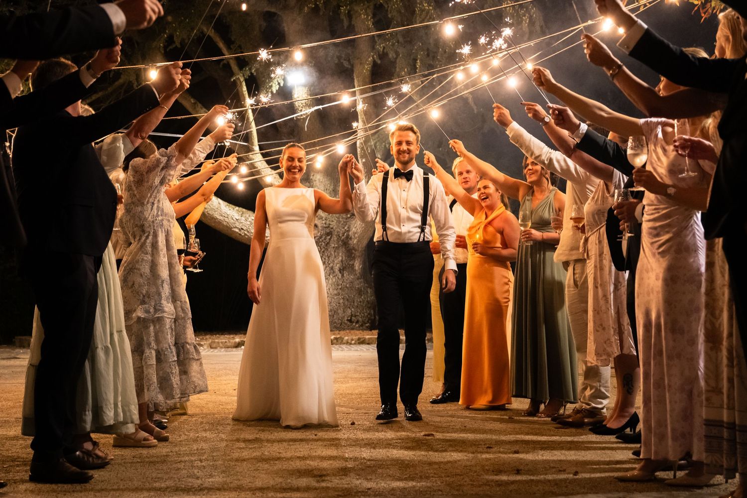 Newlyweds walk through a sparkling sendoff tunnel of guests holding lights at an outdoor evening wedding celebration.