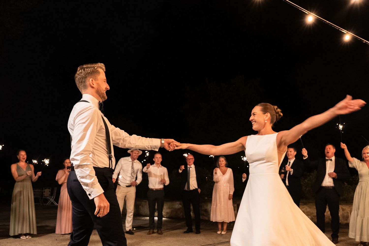 Bride and groom share first dance outdoors at night with sparklers and guests in background.