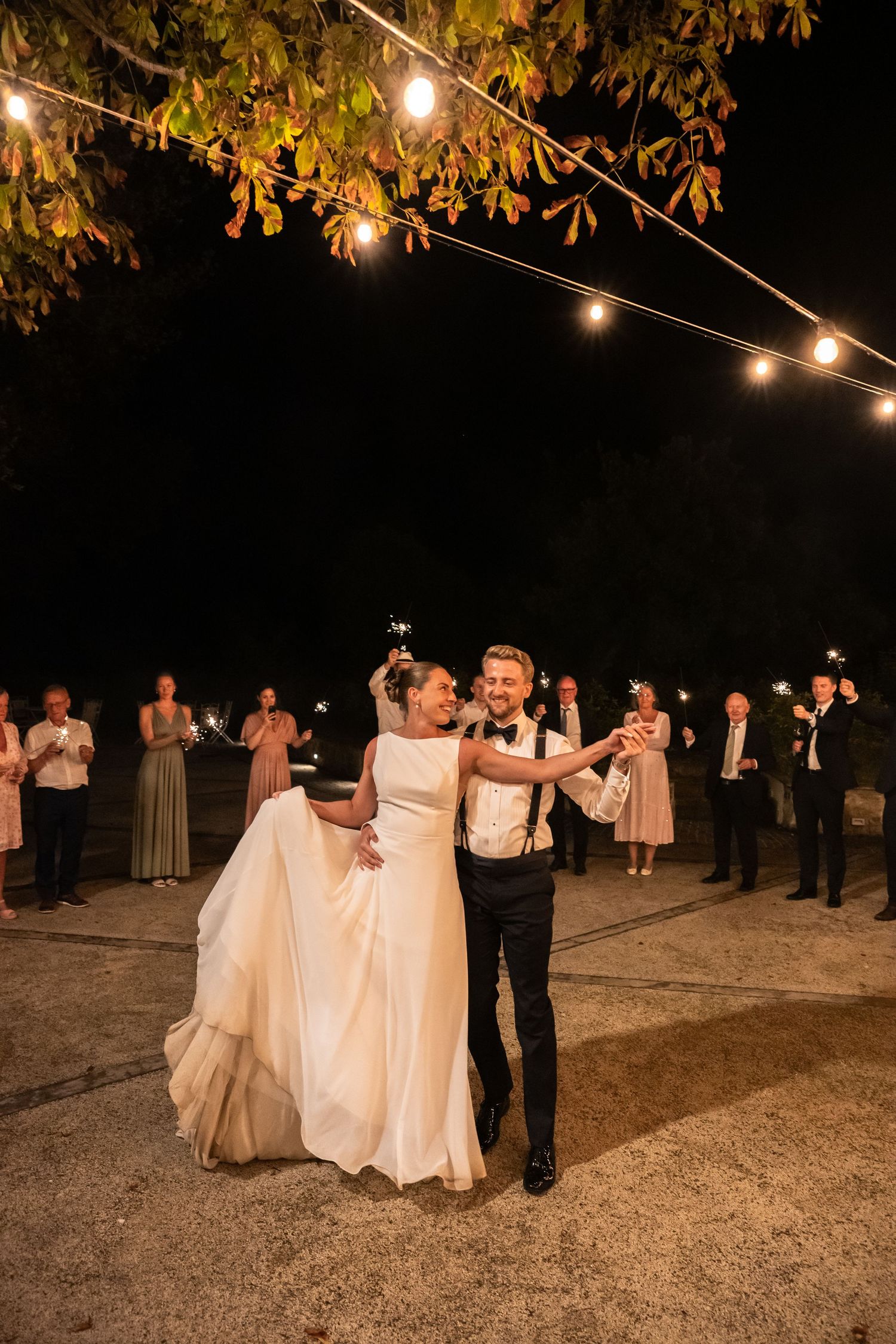 Bride and groom share first dance outdoors at night under string lights with sparkler-holding guests surrounding them.