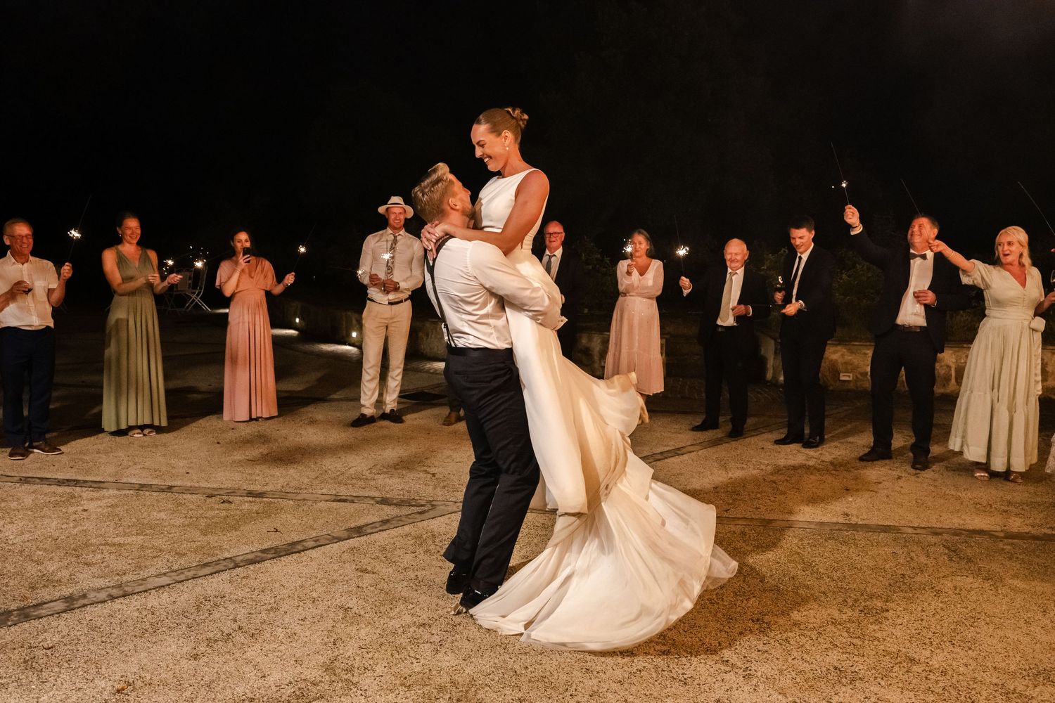Groom lifts bride in wedding dress at night outdoor celebration with sparklers and wedding party guests.