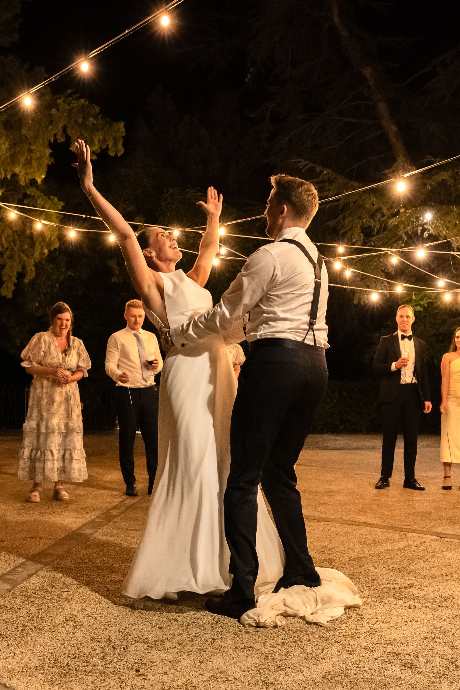 Bride and groom share first dance outdoors at night under string lights with wedding guests watching in background.