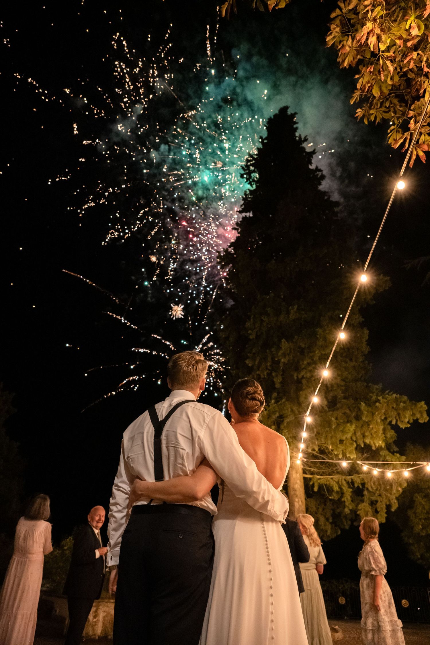 Bride and groom embrace while watching colorful wedding fireworks light up the night sky at their outdoor celebration.
