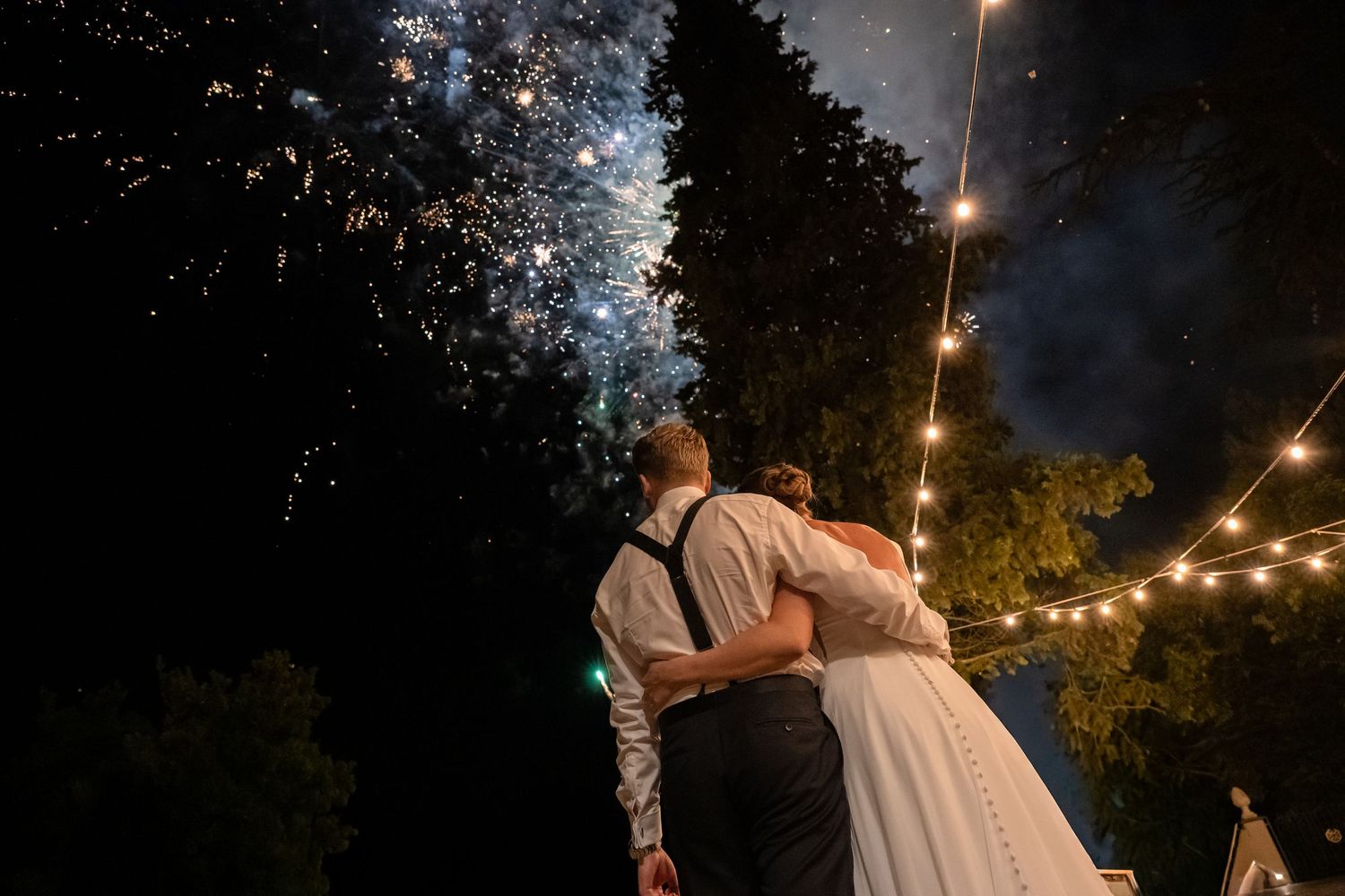 Newlyweds embrace while watching colorful fireworks display at their romantic outdoor evening wedding reception.