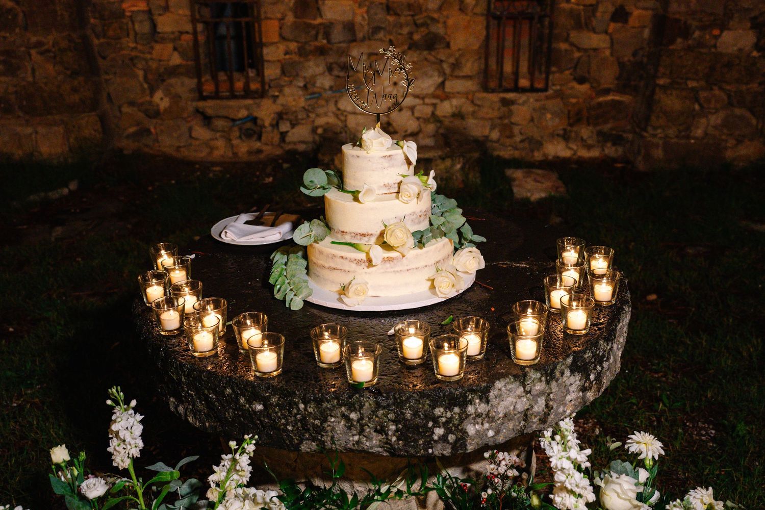 Elegant three-tier naked wedding cake surrounded by glowing candles on rustic stone table at outdoor evening reception.