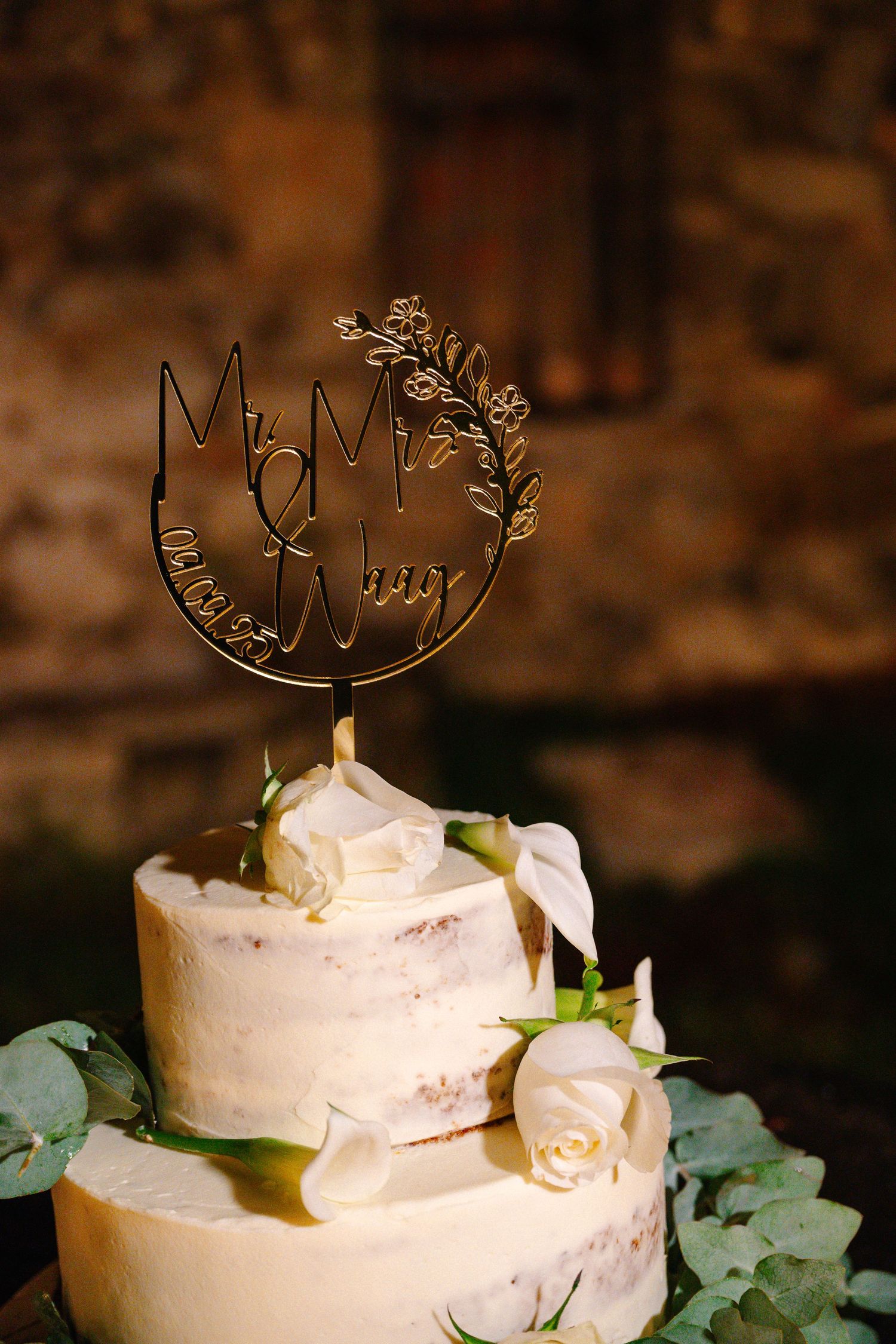 Two-tier white wedding cake with cream roses, eucalyptus, and gold Mr & Mrs cake topper on dark rustic background.