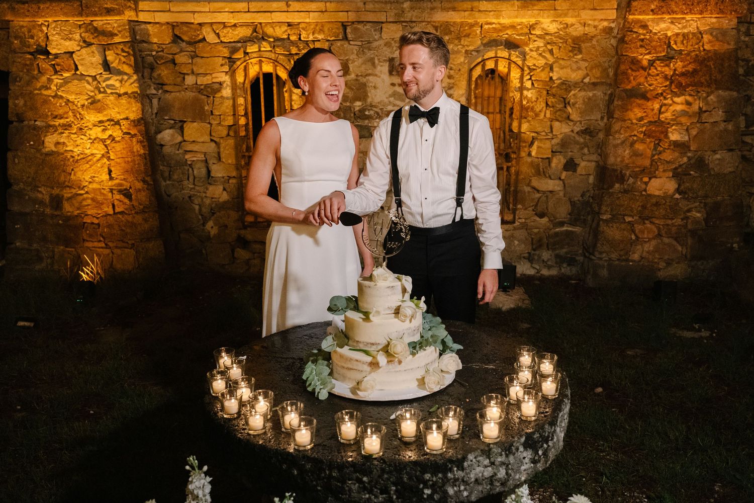 Bride and groom cutting a white wedding cake surrounded by candles at an elegant stone venue reception.
