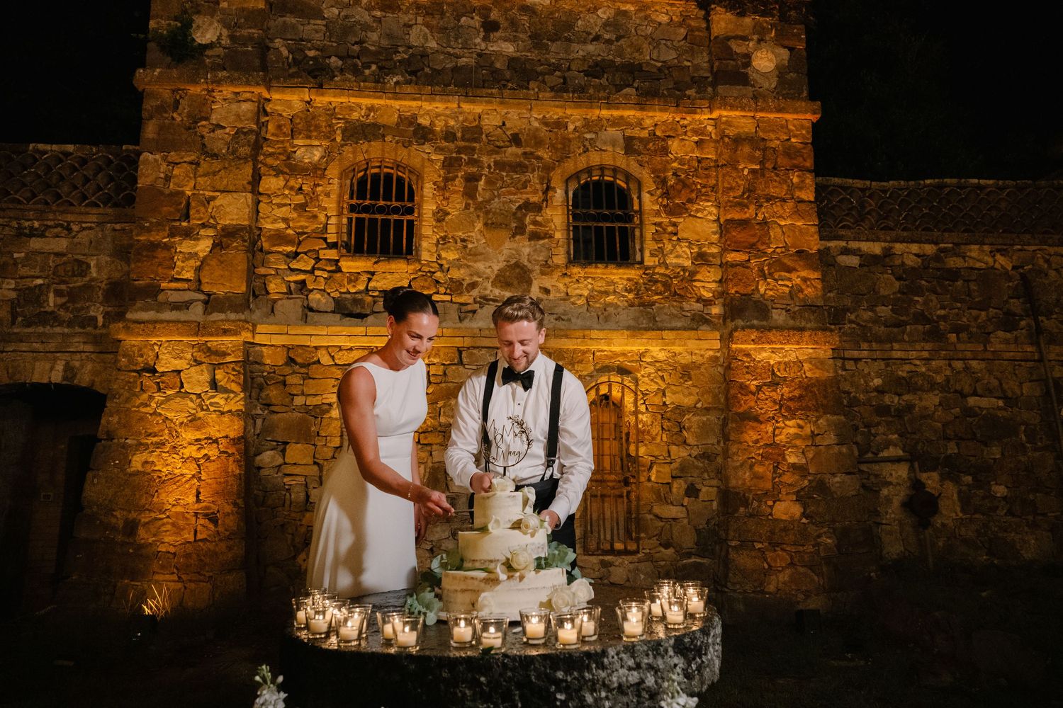 Bride and groom cutting wedding cake at night by candlelight in front of rustic stone building.