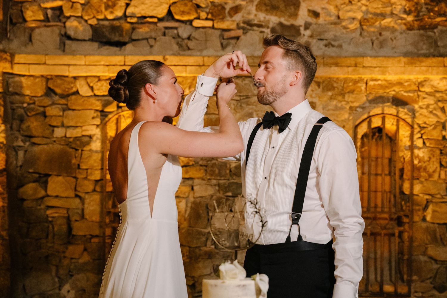 Bride feeding groom wedding cake at rustic stone wall venue, wearing white gown and suspenders.