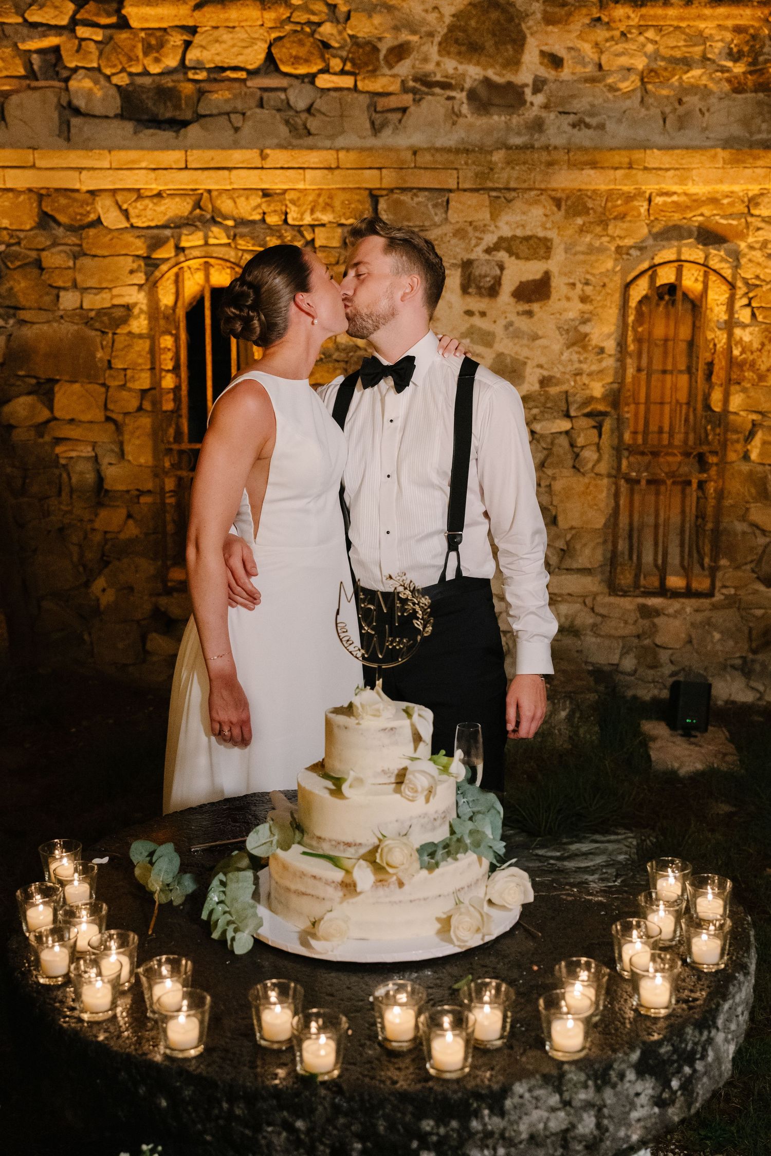 Bride and groom kissing behind a three-tier wedding cake surrounded by candles against a stone wall backdrop.