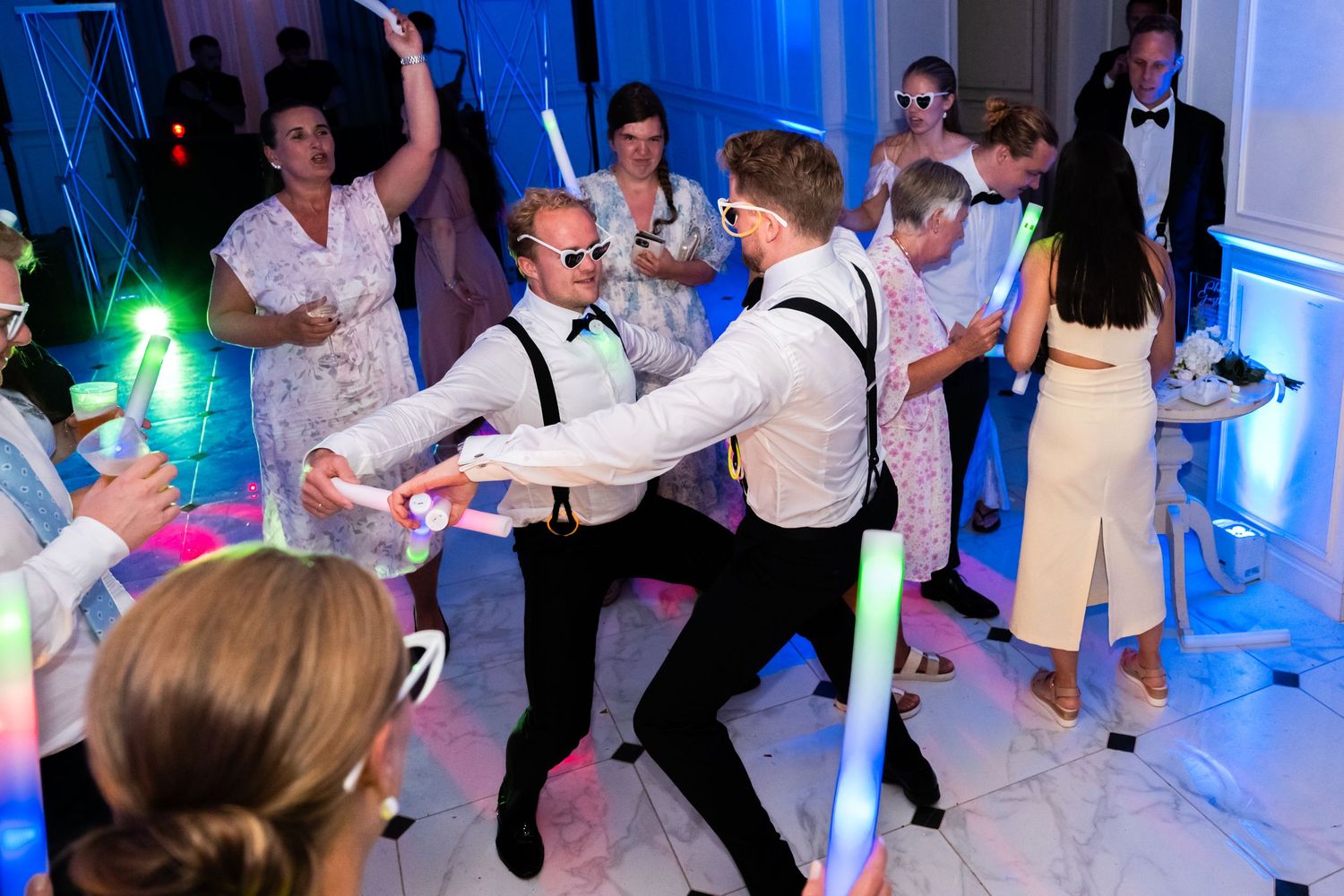 Wedding guests dancing energetically with glow sticks at a lively reception party under blue ambient lighting.