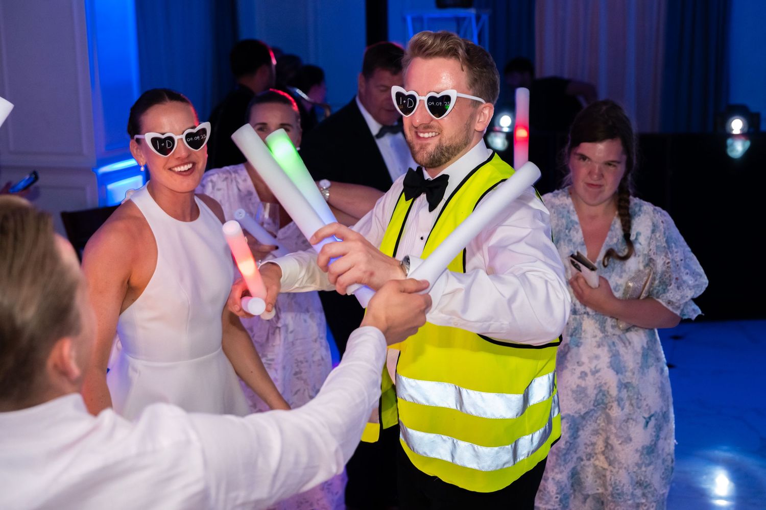 People dancing at a party wearing glow sticks, heart sunglasses, and a high-vis vest under blue and purple lights.