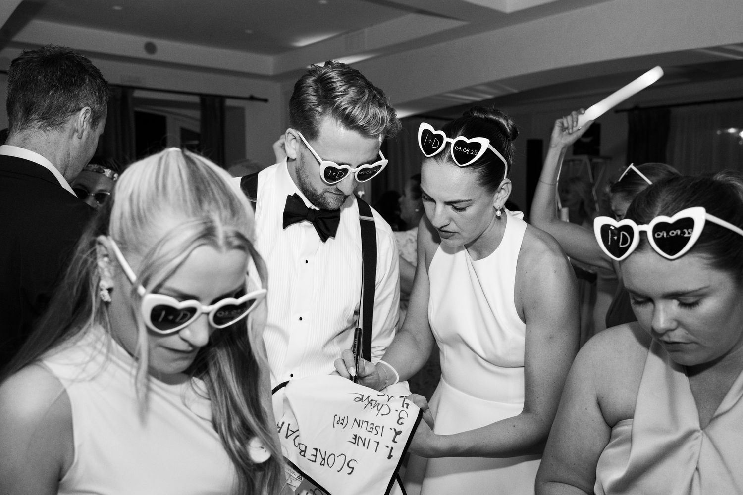 Black and white photo of wedding guests wearing heart-shaped sunglasses at a reception dance floor.