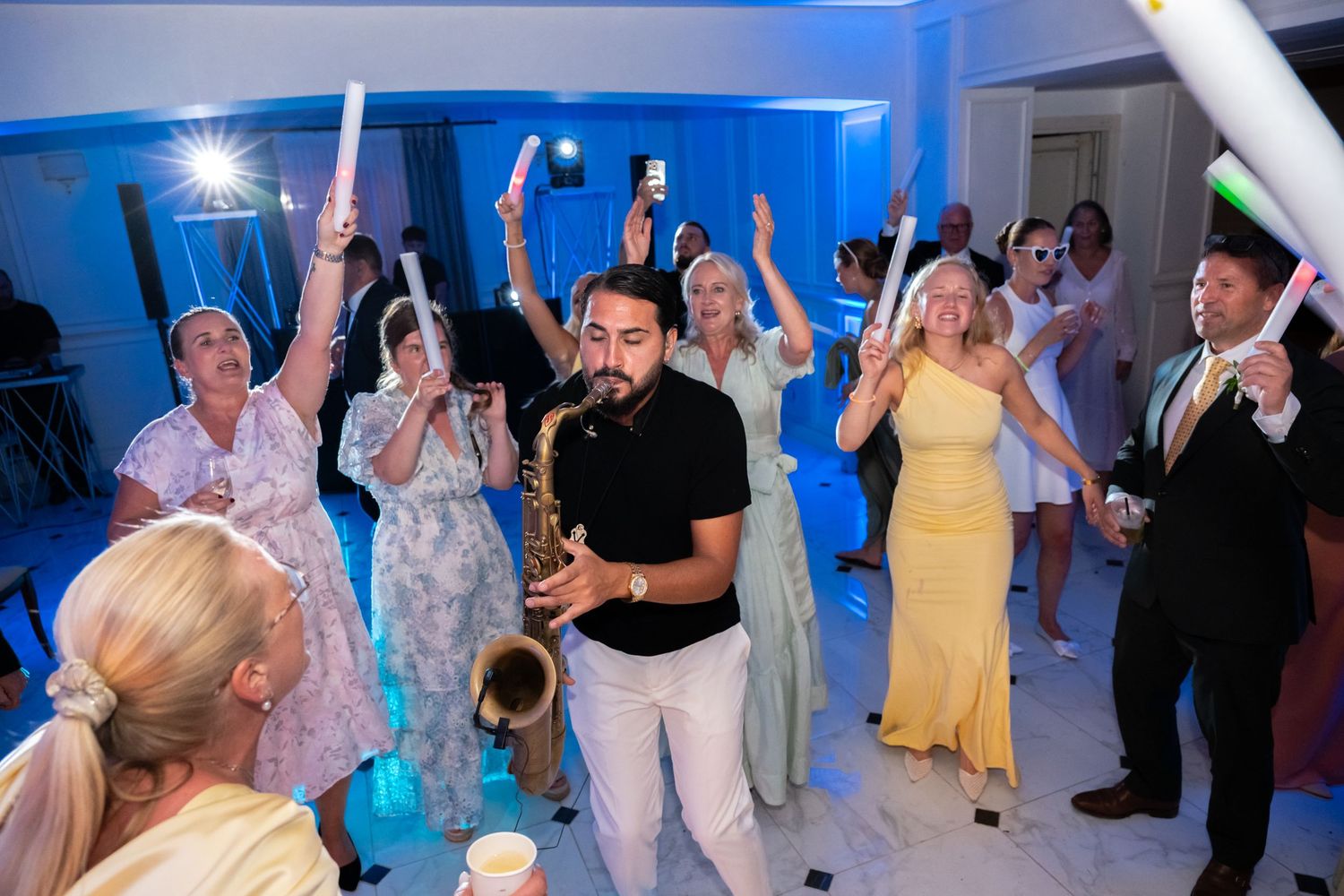 Saxophonist performs at lively wedding reception as guests dance and wave glowing foam sticks under blue lights.