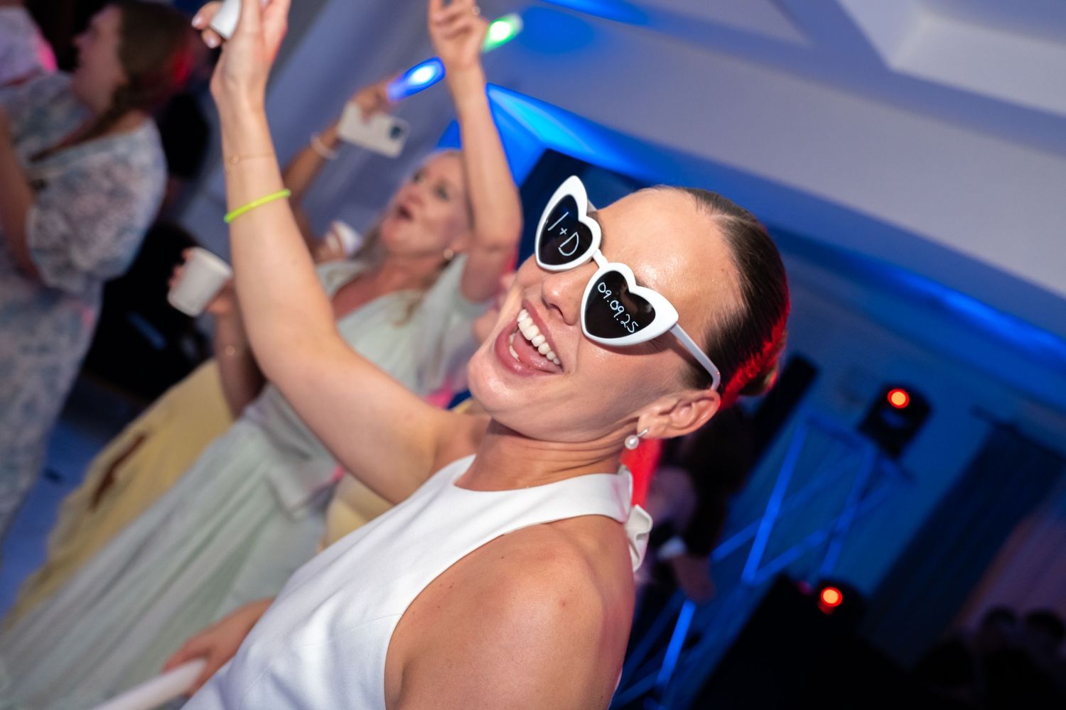 Woman in white dress and heart-shaped sunglasses dancing joyfully at a party with blue lights.