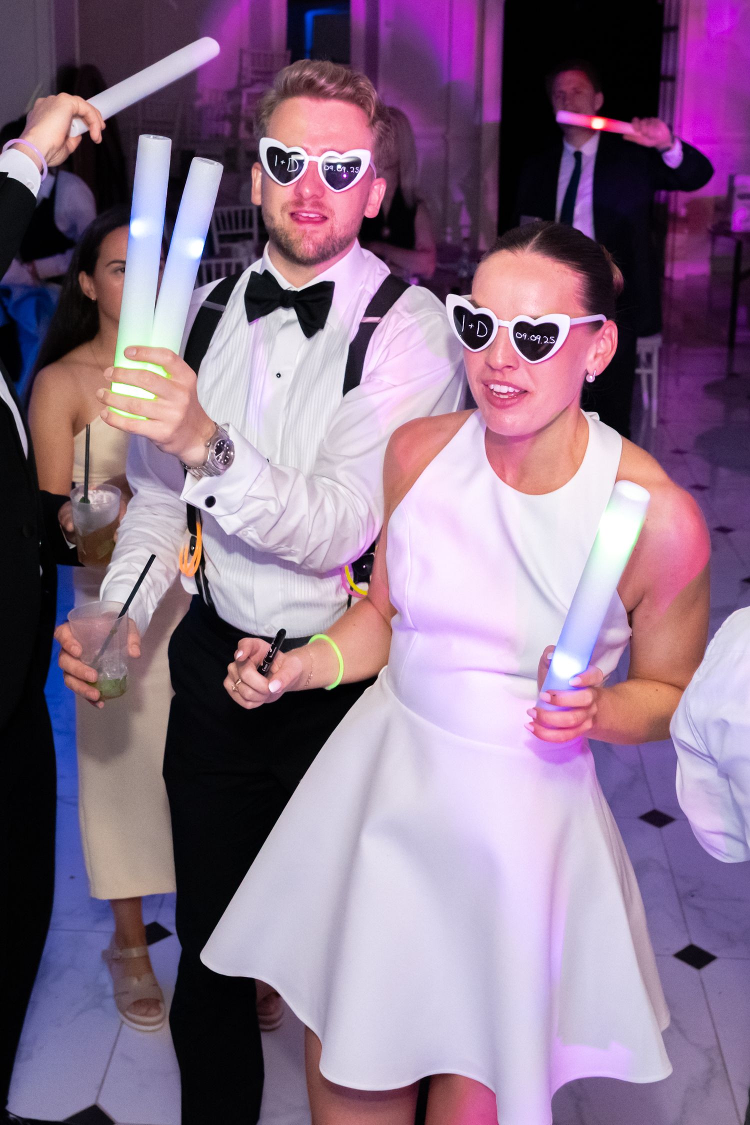 Couple in white outfits wearing heart-shaped sunglasses holding glowing foam sticks at a wedding reception party.