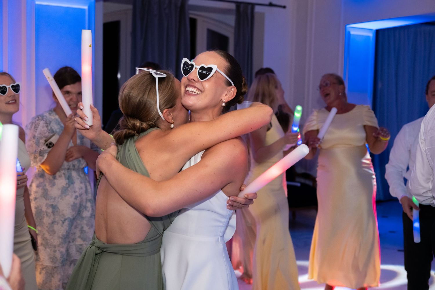 Two women hugging and dancing at a wedding reception with glowing light sticks and blue ambient lighting.