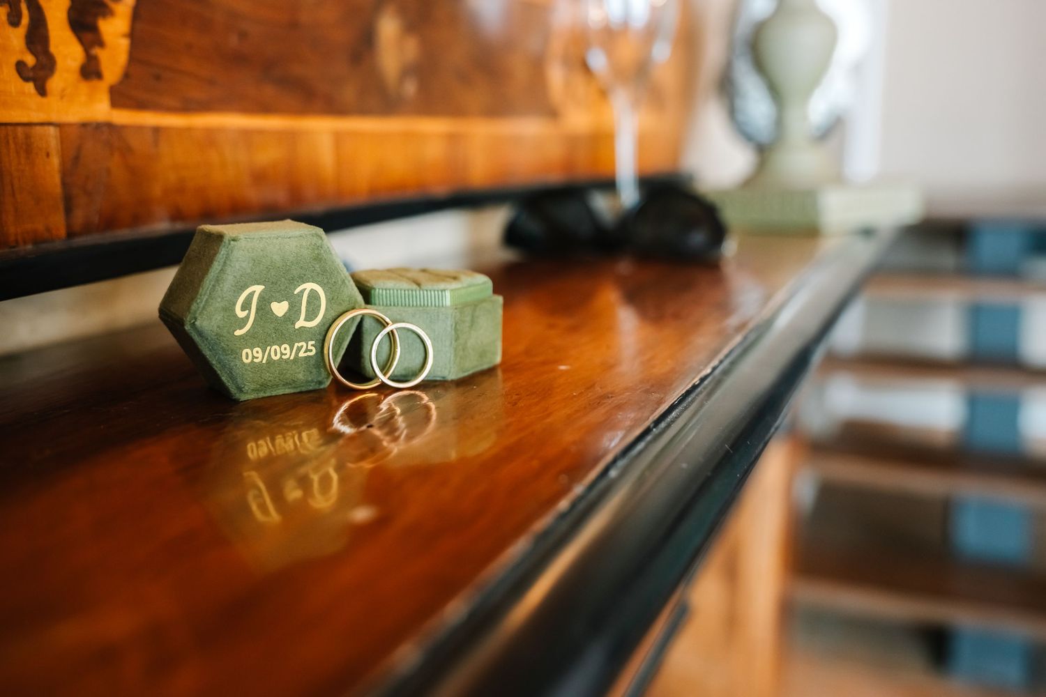 Green hexagonal wedding ring boxes with initials J and D on a polished wooden surface with gold wedding bands.