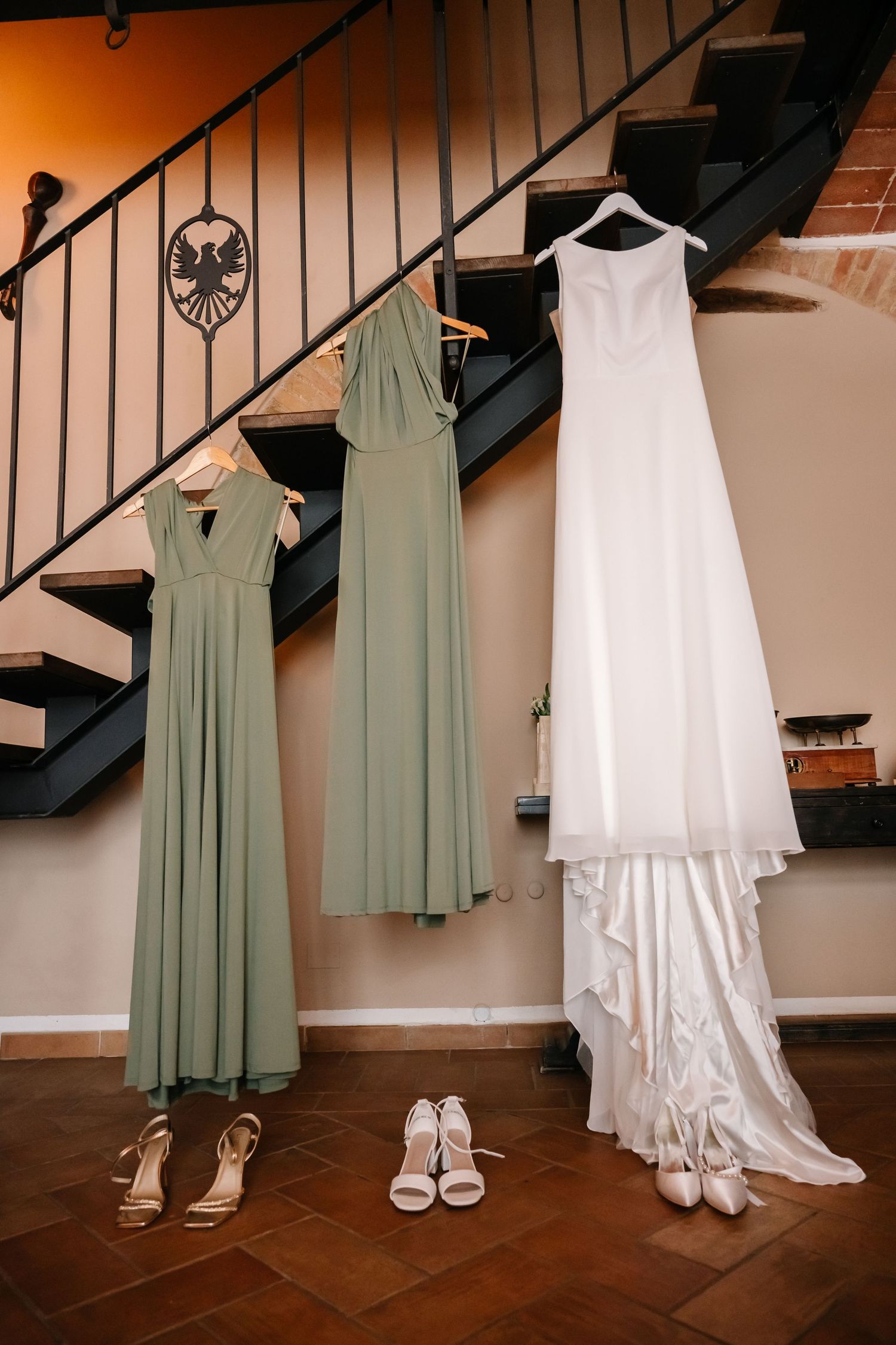 Wedding dress and sage green bridesmaid gowns hanging on staircase with heels below.